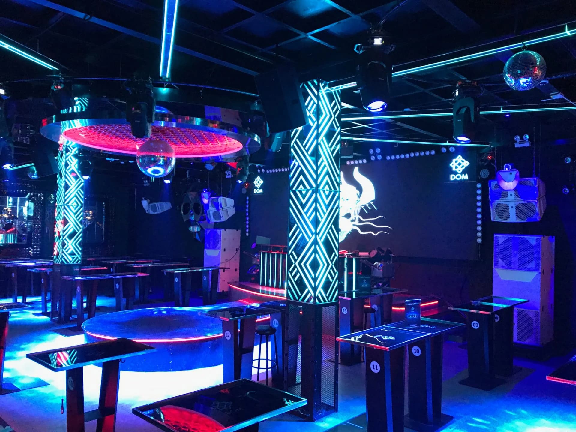 Main Floor DOM Club, TP. Nha Trang: bàn đứng đánh số quanh bục tròn, cột bọc LED, logo DOM trên màn LED và các thùng loa trắng cạnh sân khấu DJ.