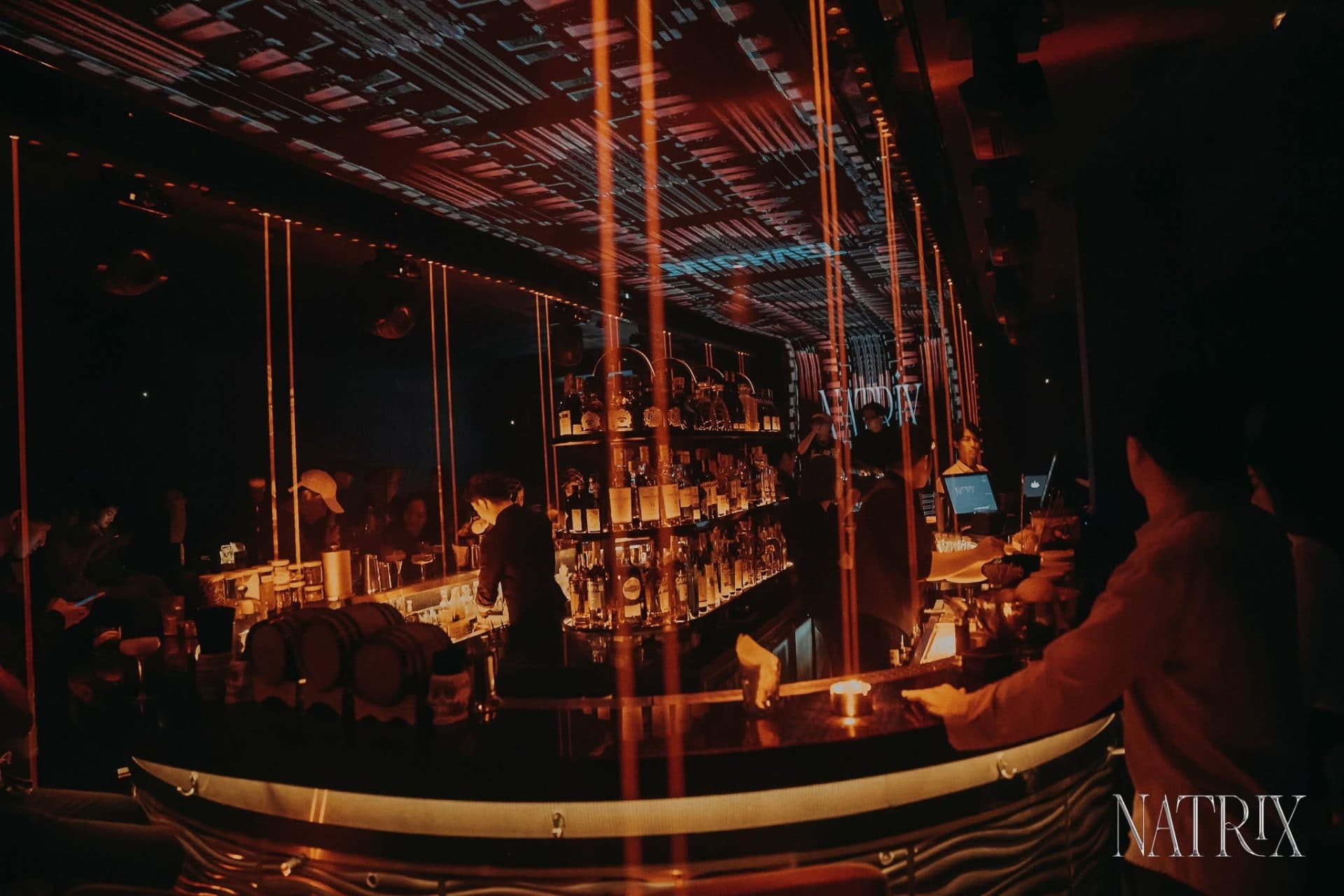Quầy bar trung tâm Natrix nhìn toàn cảnh, kệ rượu và loa gắn tường phía trên.
