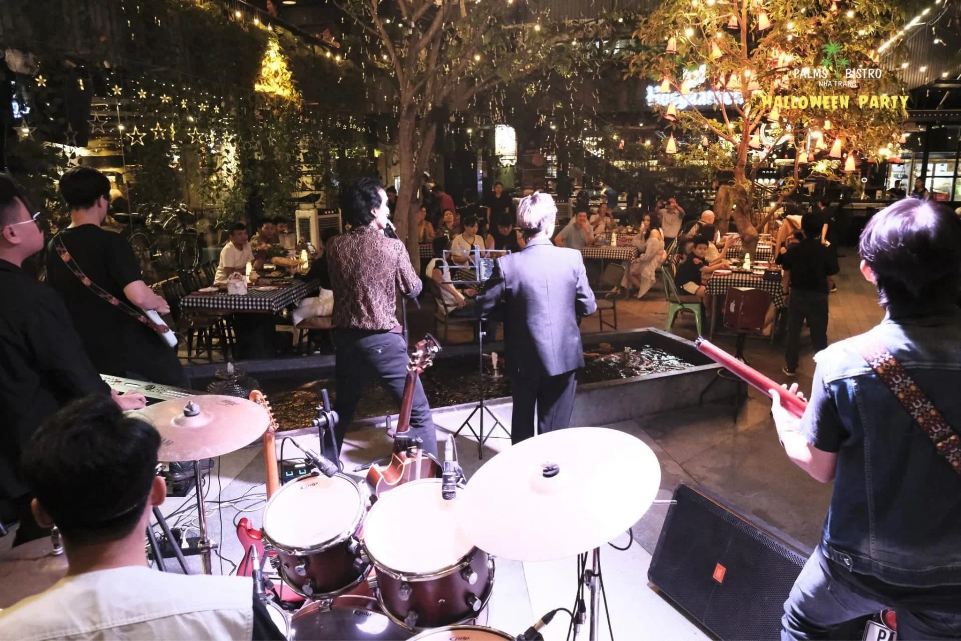 Ban nhạc biểu diễn Live trên sân khấu Palms Bistro, khán giả ngồi thưởng thức xung quanh khu vực ngoài trời.