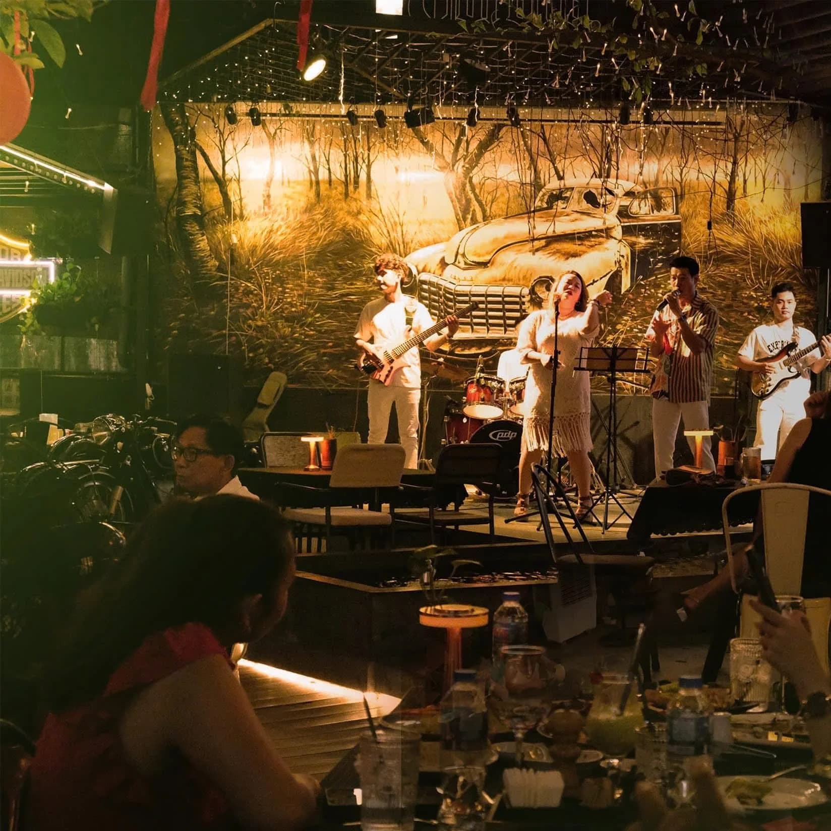 Cận cảnh ban nhạc biểu diễn buổi tối tại Palms Bistro, ánh sáng sân khấu ấm và không gian trang trí xe cổ phía sau.
