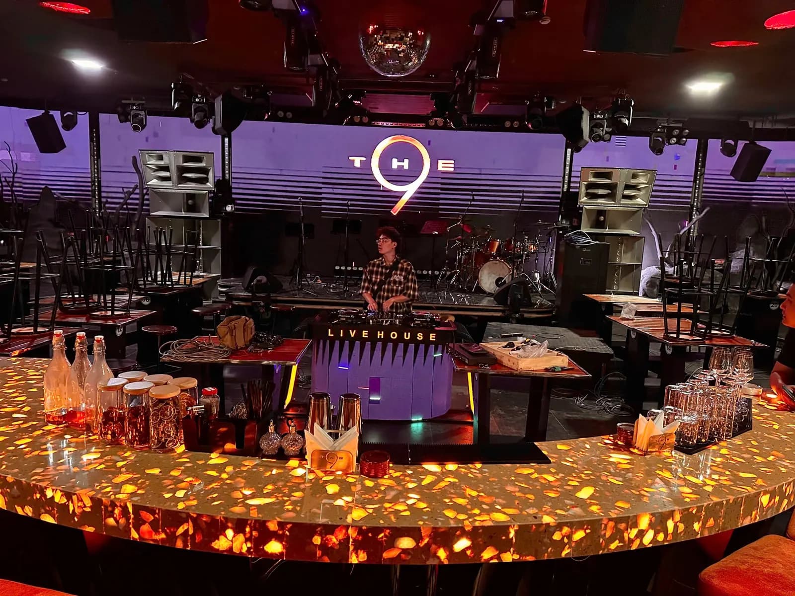 Toàn cảnh The 9 nhìn từ quầy bar — hai cụm Main PA treo đối xứng hai bên sân khấu, bục DJ LIVE HOUSE và bộ trống Live Band trước màn hình LED.