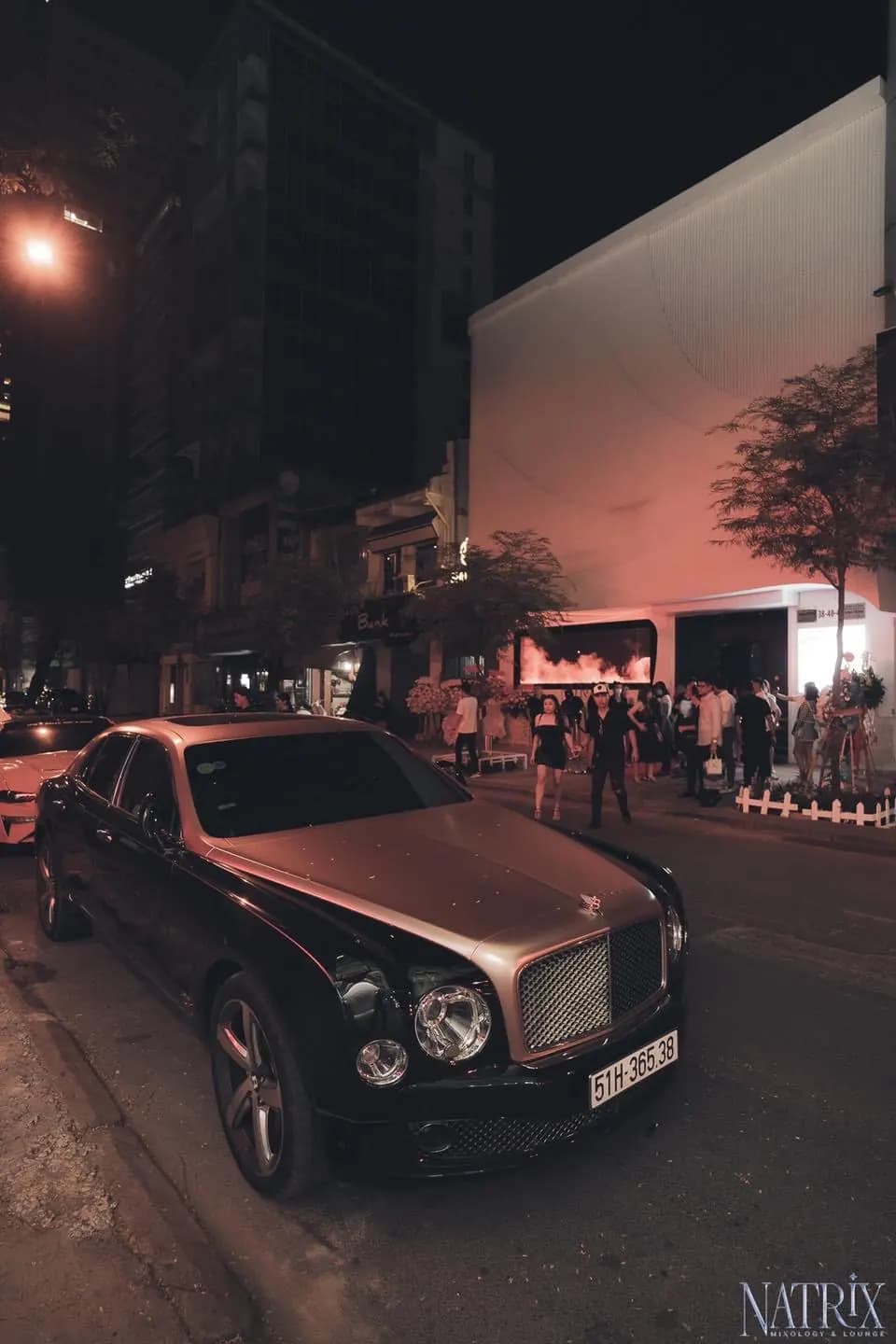 Mặt tiền Natrix Bar về đêm nhìn từ vỉa hè, xe hơi đỗ trước quán và khách đứng trò chuyện.