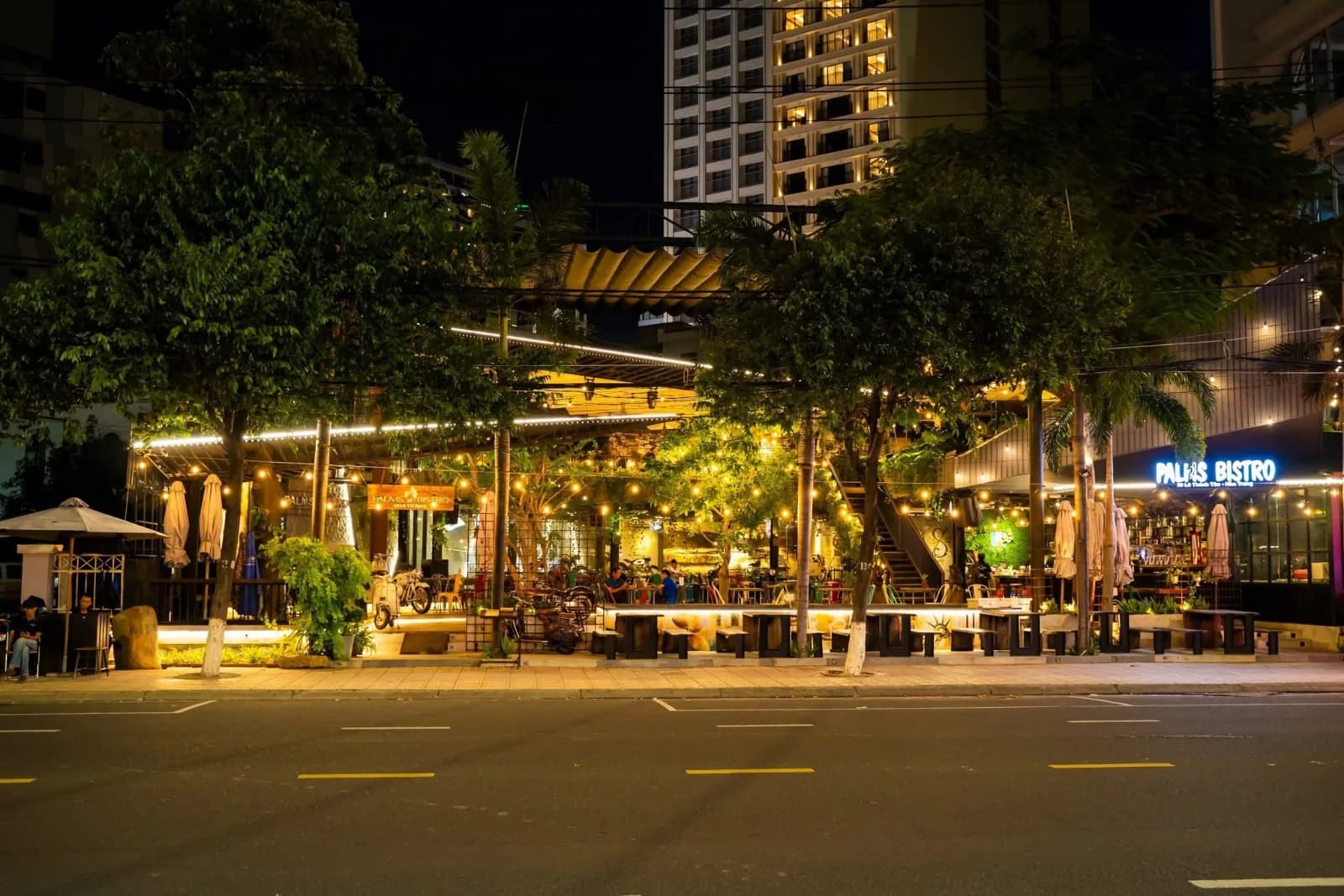Palms Bistro về đêm nhìn từ bên kia đường Lê Thánh Tôn, TP. Nha Trang: mặt tiền mở dưới dây đèn, cây xanh và dãy bàn ra sát vỉa hè.