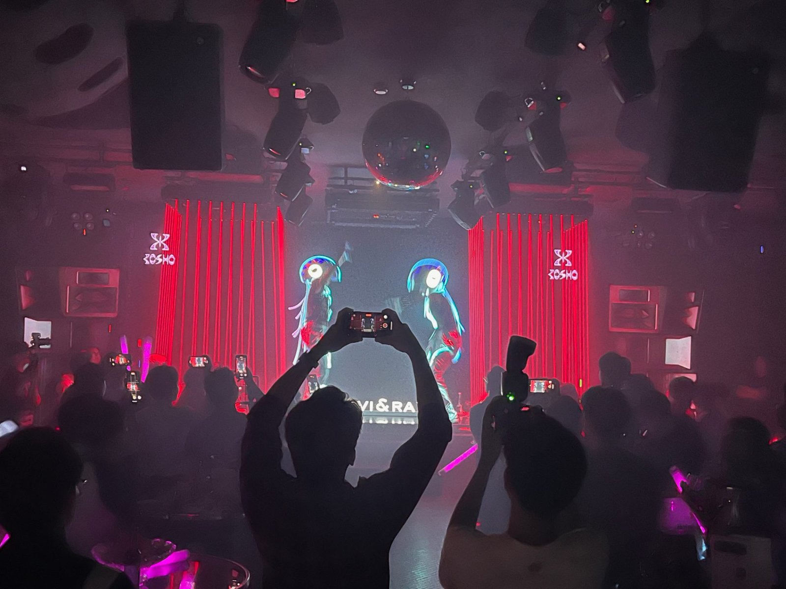 Đám đông khán giả giơ điện thoại quay lại sân khấu Kosho Club dưới ánh đèn đỏ, hai vũ công LED trình diễn giữa dải rèm đỏ và bảng hiệu neon Kosho hai bên.