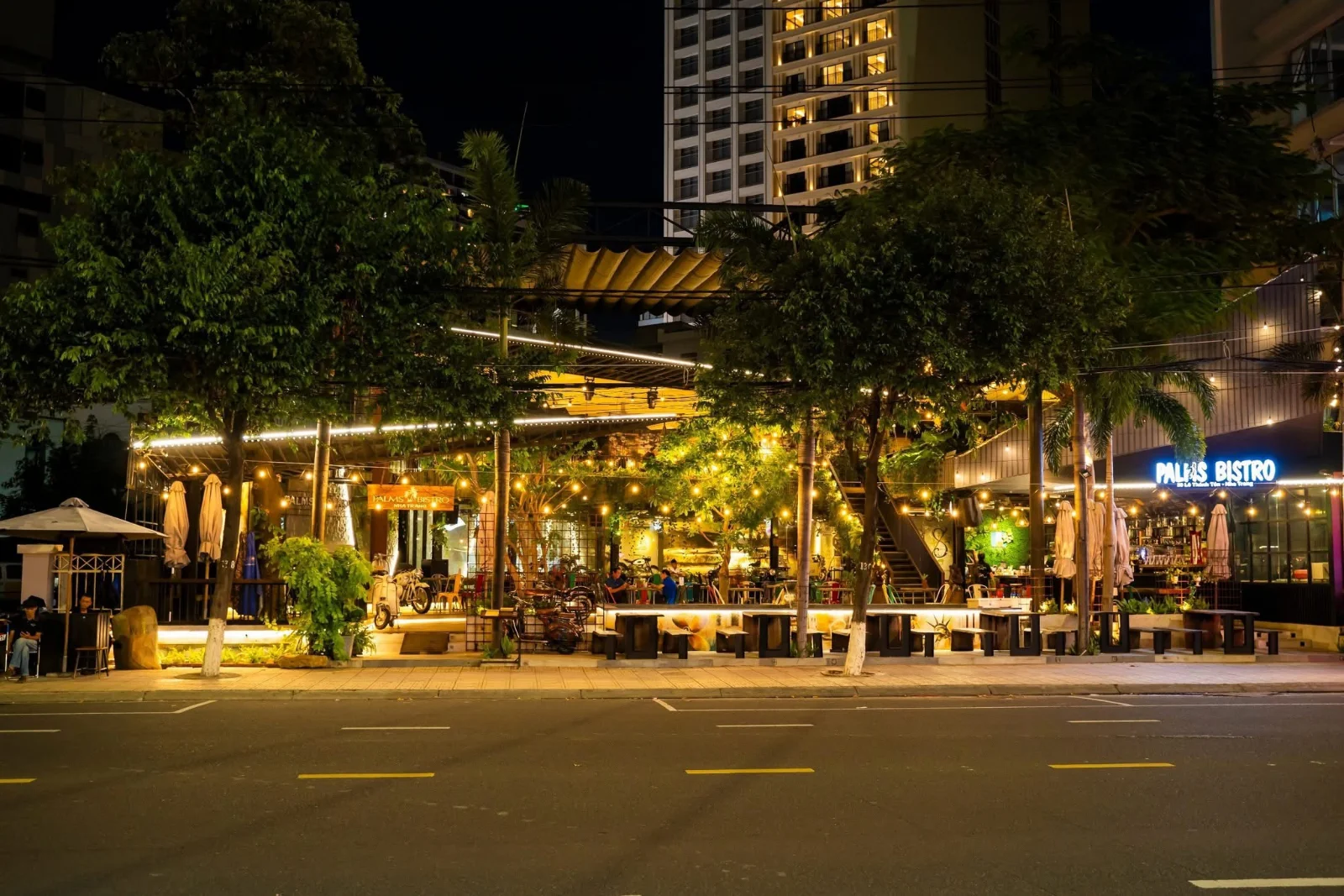 Palms Bistro về đêm nhìn từ bên kia đường Lê Thánh Tôn, TP. Nha Trang: mặt tiền mở dưới dây đèn, cây xanh và dãy bàn ra sát vỉa hè.